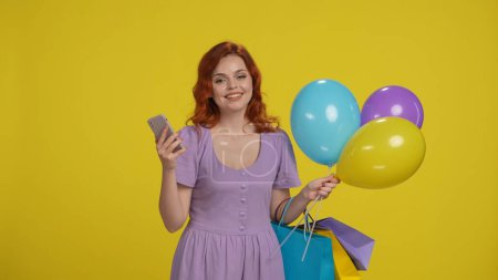 Téléchargez les photos : Femme avec sacs à provisions et ballons en studio sur fond jaune. Une femme utilise un smartphone - en image libre de droit
