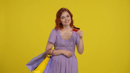 Foto de Una mujer con bolsas de compras tiene una tarjeta de crédito con una sonrisa. Mujer pelirroja en un vestido de noche en el estudio sobre un fondo amarillo - Imagen libre de derechos