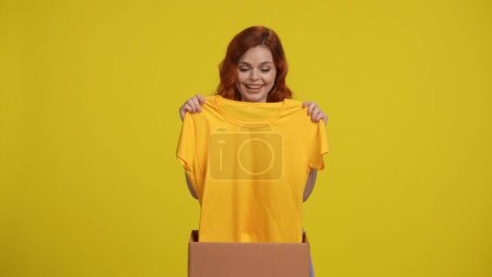 Téléchargez les photos : Achats rapides en boutique en ligne. Une femme déballe une boîte livrée d'une boutique en ligne et examine avec enthousiasme un nouveau T-shirt. Femme en studio sur fond jaune. Concept de livraison - en image libre de droit