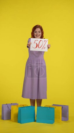 Photo for An attractive young woman stands surrounded by shopping bags and holds a sign that says fifty percent. A redhaired woman stands full length in a studio against a yellow background. Vertical shot - Royalty Free Image