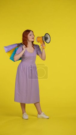 Photo for Happy redhaired woman with shopping bags speaking into a megaphone. Full length woman in the studio on a yellow background. Vertical shot. Sale and discount concept - Royalty Free Image