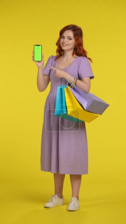 Photo for A charming redhaired woman points her index finger at the green screen of a smartphone in her hand. A woman with shopping bags in full length in studio on yellow background. Vertical shot. Black - Royalty Free Image