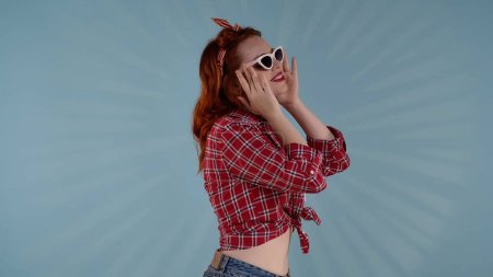 Téléchargez les photos : Dans le cadre, une jeune femme aux cheveux roux avec un maquillage lumineux se tient sur le côté de la caméra sur un fond bleu. Elle porte des lunettes de soleil et regarde quelque part. Un tir moyen. Matériel HDR BT2020 HLG - en image libre de droit