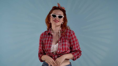 Photo for In the frame against a blue background is a young red-haired woman with brightly colored makeup. She is standing in sunglasses facing the camera and looking somewhere. Medium shot. HDR BT2020 HLG - Royalty Free Image