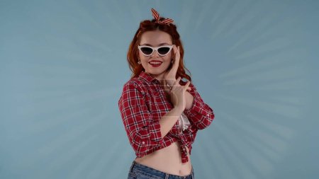 Téléchargez les photos : Dans le cadre, une jeune femme aux cheveux roux avec un maquillage lumineux se tient sur le côté de la caméra sur un fond bleu. Elle porte des lunettes de soleil et regarde la caméra. Un tir moyen. HDR BT2020 HLG - en image libre de droit