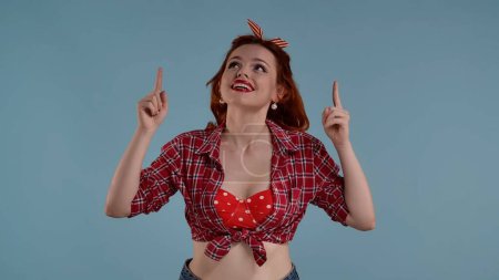 Photo for A young red-haired girl with brightly colored makeup is in the frame against a blue background. She looks and points upward with her index fingers on both hands. Here could be your advertisement. HDR - Royalty Free Image