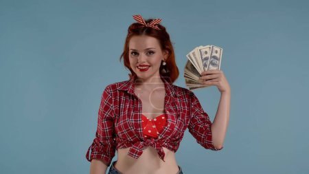 Photo for Framed against a blue background is a young red-haired woman with brightly colored makeup. Holding a fan of money in her hands. She is looking at the camera and as if waving it. Demonstrates pleasure - Royalty Free Image