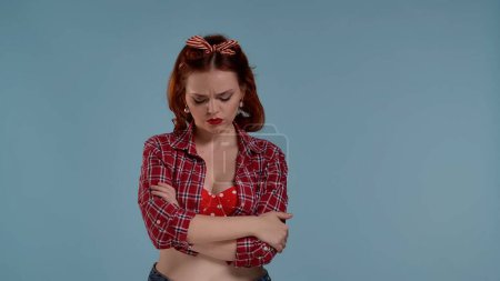 Photo for Framed against a blue background is a young red-haired woman with brightly colored makeup. Folding her arms and lowering her eyes down the woman is sad. Perhaps someone has wronged her, she is sad - Royalty Free Image