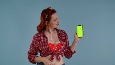 Foto de En el marco sobre un fondo azul joven mujer pelirroja con maquillaje brillante. Mira el teléfono, que tiene una pantalla verde. Expresa sorpresa, alegría. Aquí puede ser su anuncio, producto. HDR - Imagen libre de derechos