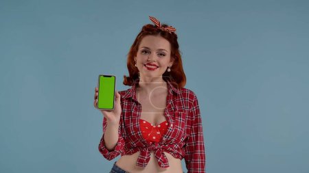 Photo for In the frame on a blue background young woman. Looks at the camera and smiles. Holding a cell phone with a green screen in front of her. This could be your advertisement, your product. Medium shot - Royalty Free Image