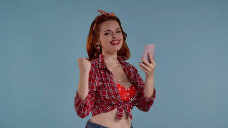 Téléchargez les photos : La photo montre une jeune femme aux cheveux roux avec un maquillage de couleur vive sur un fond bleu. Elle regarde dans la caméra tenant un téléphone portable. La surprise, la joie et le bonheur sont également représentés. HDR - en image libre de droit