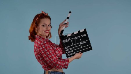In the frame on a blue background is a young woman with brightly colored makeup. She is looking at the camera, smiling and holding a clapper in her hands. Demonstrates filming a movie, TV series. HDR