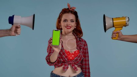 Photo for Young redheaded woman. She looks at the camera and smiles. In front of her, she holds a phone with a green screen. On both sides she is pointed loudspeakers, horn. Here may be your advertisement. HDR - Royalty Free Image