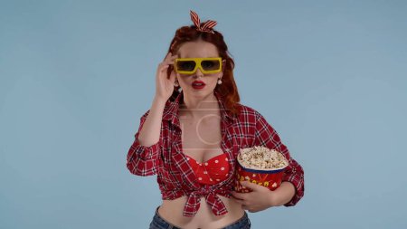 Téléchargez les photos : Dans le cadre sur un fond bleu se trouve une jeune femme rousse. Elle regarde la caméra en lunettes 3D, mangeant du pop-corn. Démontre regarder un film dans un théâtre, dépeint la peur, l'inconfort. HDR - en image libre de droit
