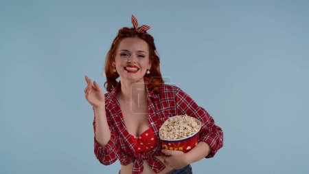 Photo for In the frame on the blue background is a young red-haired woman with bright makeup. She looks at the camera and tosses popcorn. Demonstrates watching a movie in a theater she laughs. Joyful, happy - Royalty Free Image