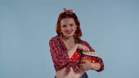 Photo for In the frame on the blue background is a young red-haired woman with bright makeup. She is looking at the camera and eating popcorn. Demonstrating watching a movie in a theater she smiles. Joyful. HDR - Royalty Free Image