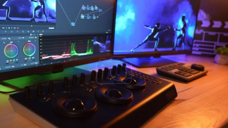 Photo for Video and photo editing advertisement concept. Freelancer workspace at home. Closeup shot of monitors, editing equipment for video on the table, screen shows software setup, neon light at the back. - Royalty Free Image