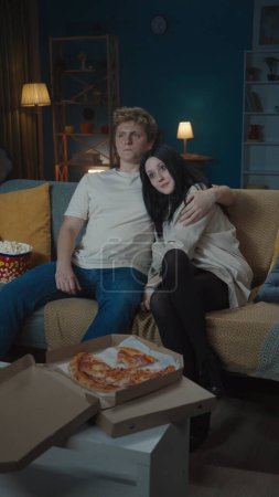 Photo for First relationships creative concept. Portrait of teenage couple spending leisure time. Boy and girl sitting on the sofa eating popcorn and watching tv and hugging. Vertical photo. - Royalty Free Image