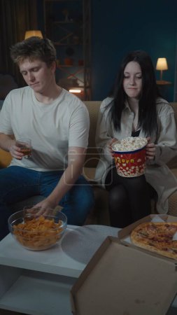 Photo for First relationships creative concept. Portrait of teenage couple spending leisure time. Boy and girl sitting on the sofa. Boy holds glasses with whiskey girl looks negative. Vertical photo. - Royalty Free Image