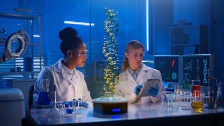 Téléchargez les photos : Photo de taille moyenne capturant deux femmes scientifiques portant des lunettes et des blouses de laboratoire assises dans un laboratoire, faisant des recherches sur l'hologramme de l'ADN affiché devant elles. Avancé, innovation, réalité augmentée, science - en image libre de droit