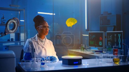 Photo for Medium-sized photo capturing a dark-skinned female scientist wearing glasses and a lab coat sitting in a laboratory, researching brain hologram. Advanced, innovation, augmented reality, science. - Royalty Free Image