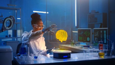 Mittelgroßes Foto, auf dem eine dunkelhäutige Wissenschaftlerin mit Brille und Labormantel in einem Labor sitzt und das Hologramm des Gehirns erforscht. Fortschrittlichkeit, Innovation, erweiterte Realität, Wissenschaft.