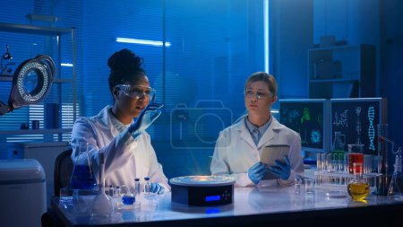 Téléchargez les photos : Photo de taille moyenne capturant deux femmes scientifiques portant des lunettes et des blouses de laboratoire assises dans un laboratoire, faisant des recherches sur votre hologramme affiché devant elles. Réalité augmentée, science, modèle, maquette. - en image libre de droit