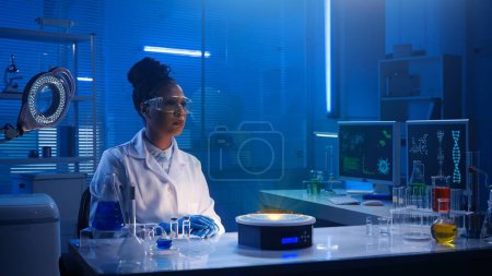 Photo for Medium-sized photo capturing a female scientist wearing glasses and lab coats sitting in a lab, researching your hologram displayed in front of them. Augmented reality, science, template, mock up. - Royalty Free Image
