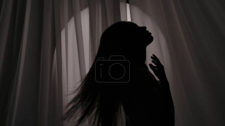 Foto de Foto mediana de una silueta de mujer joven envuelta en una toalla tocando, acariciando su cuerpo, cuello y agitando su cabello en una luz amortiguada. Anuncio de productos, rutina de autocuidado. - Imagen libre de derechos