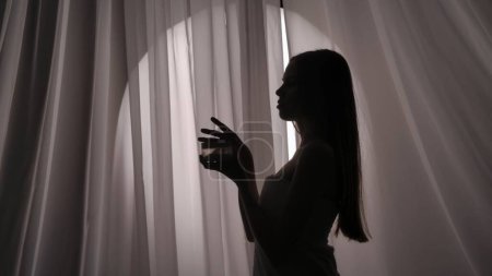 Foto de Vista lateral media de una silueta de mujer joven envuelta en una toalla sosteniendo un frasco de cuerpo, crema facial, producto de belleza en una luz amortiguada. Anuncio de productos, rutina de autocuidado. - Imagen libre de derechos