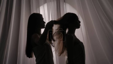 Photo for Medium side view shot of two sisters, friends, young womens silhouettes wrapped in a towel. One of them is braiding the other ones hair in a muffled light. Product advertisement, self-care routine. - Royalty Free Image