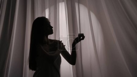Foto de Foto de vista lateral media de una silueta de mujer joven envuelta en una toalla sosteniendo un frasco de cuerpo, crema facial y aplicando la sustancia sobre su piel en una luz amortiguada. Publicidad de productos, autocuidado. - Imagen libre de derechos