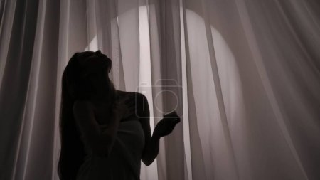 Foto de Foto de vista lateral media de una silueta de mujer joven envuelta en una toalla sosteniendo un frasco de cuerpo, crema facial y aplicando la sustancia sobre su piel en una luz amortiguada. Publicidad de productos, autocuidado. - Imagen libre de derechos