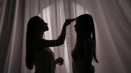 Photo for Medium side view shot of two sisters, young womens silhouettes wrapped in a towel. One of them is caressing, brushing the other ones hair in a muffled light. Product advertisement, care routine. - Royalty Free Image