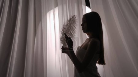 Foto de Foto de vista lateral media de una silueta de mujer joven envuelta en una toalla con una pluma blanca en la mano, tocándola suavemente. Producto, ocio, publicidad de servicios, rutina de autocuidado. - Imagen libre de derechos