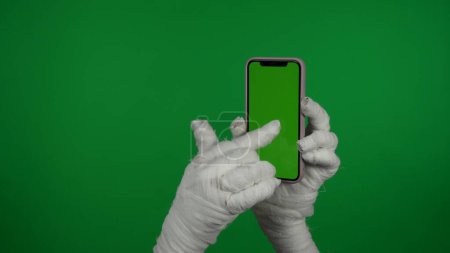 Téléchargez les photos : Détail écran vert isolé chroma photo clé d'une momie main tenant un smartphone avec une zone publicitaire, espace de travail maquette pour votre clip de promotion et tapant sur l'écran. - en image libre de droit