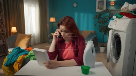 Photo for Redhead woman sitting next to a washing machine, talking on the phone with somebody so they could help in choosing between powdered and liquid detergents. Household appliances, chores, advertisement. - Royalty Free Image
