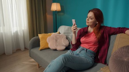 Photo for Full-size shot capturing a young redhead woman sitting on the couch, chatting with somebody, surfing online, scrolling his feed in social network. Indoor, media, blogger, hobbies and leisure. - Royalty Free Image