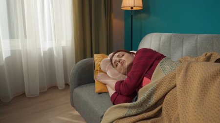Téléchargez les photos : Femme rousse dormant, faisant la sieste sur le canapé, canapé dans le salon, posant la tête sur l'oreiller et recouvert d'une couverture, se sentant confortable. Épuisement, détente, temps libre, loisirs. - en image libre de droit