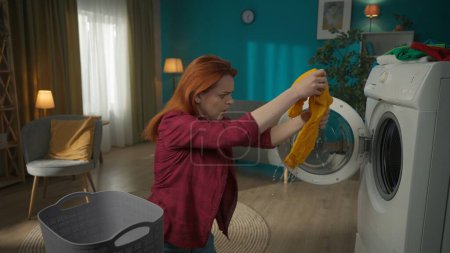 Photo for Redhead woman standing next to a washing machine, unloading it. The clothes are soaking wet, the woman is stressed that machine didnt complete the drying. Household appliances, chores, advertisement. - Royalty Free Image