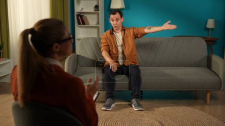 Photo for The indignant man points with his hand in the direction his wife has gone. The joint psychotherapy session has failed. A man sitting on a couch in front of a female psychologist - Royalty Free Image