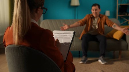 Photo for Rear view of a female psychologist against a blurred background of a male patient sitting on a couch in front of her. The therapist is filling out a diagnostic sheet while talking to the male patient - Royalty Free Image
