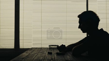 Foto de El video muestra a un joven sentado en una mesa en un café. Se sienta solo con cigarrillos y un cenicero sobre la mesa. Está a punto de fumar y demuestra soledad, adicción. Tiro medio. - Imagen libre de derechos