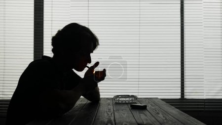 Foto de En una silueta, un joven se sienta en una mesa en un café. Hay cigarrillos y un cenicero sobre la mesa. Enciende un cigarrillo para fumar. Demuestra adicción, soledad, tristeza. Tiro medio. - Imagen libre de derechos