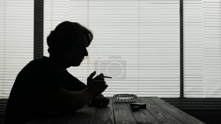 Foto de En la silueta de un joven sentado en una mesa en un café. Fuma un cigarrillo, suelta humo y mira en alguna parte. Demuestra adicción, soledad, tristeza. Tiro medio. - Imagen libre de derechos