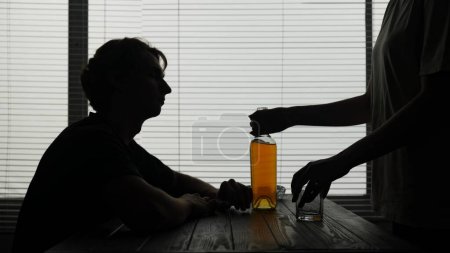 Photo for In the frame, a young guy sitting at a table in an institution, a cafe. He is brought a bottle of alcohol apparently by a waiter and a glass. Hes going to drink it alone. Alcohol addiction, sadness. - Royalty Free Image