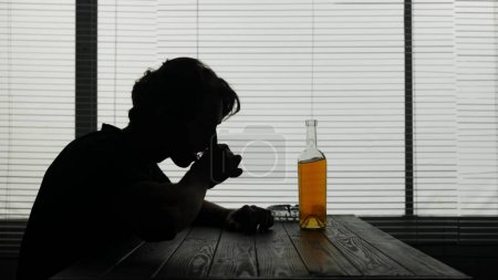 Photo pour Dans le cadre, un jeune homme est assis dans un café. Il est frustré, triste. Puis prend une bouteille le verse dans un verre et boit l'alcool. Démontre une dépendance à l'alcool, la solitude, la tristesse. - image libre de droit