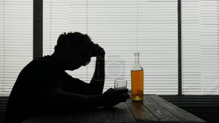 Photo for In the silhouette of a young man sitting at a table in a cafe. Nearby is a bottle of alcohol in his hands he holds a glass and thinks about something. He shows alcoholism, sadness and loneliness. - Royalty Free Image
