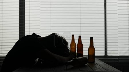 Photo for In the silhouette of a young man sitting at a table in a cafe. Nearby are empty bottles of alcohol. He demonstrates alcohol intoxication, so he fell asleep at the table. He is lonely and dependent. - Royalty Free Image