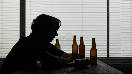 Téléchargez les photos : Dans la silhouette d'un jeune homme assis à une table dans un café. A proximité se trouvent des bouteilles d'alcool vides. Il démontre une intoxication alcoolique assis seul il est triste et accro. Plan moyen. - en image libre de droit